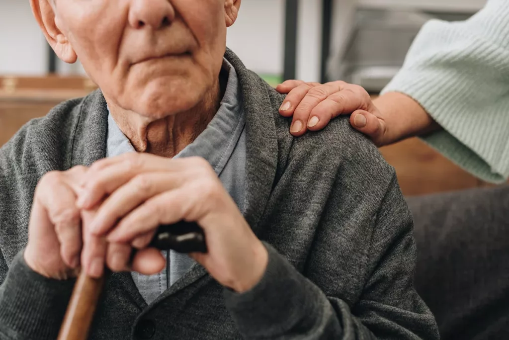 cropped view of senior husband sitting in living room with senior wife lovingly resting her hand on his shoulder, symbolizing struggles of dementia 